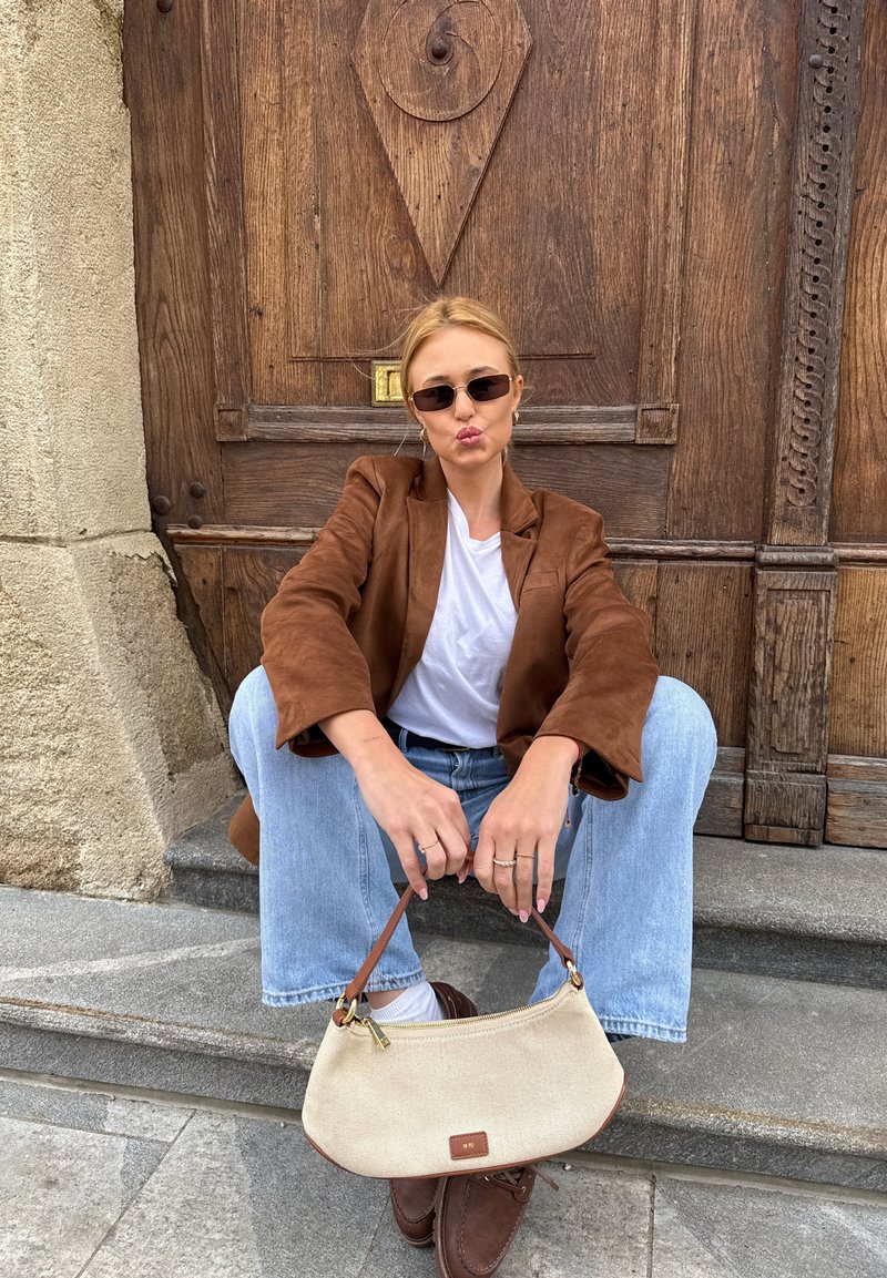 Junge Frau mit Sonnenbrille, brauner Jacke, weißem Hemd und hellblauen Jeans sitzt auf Steinstufen und hält eine cremefarbene Handtasche vor einer verzierten Holztür.