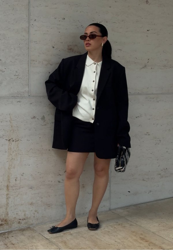 Vrouw met zwarte blazer en rok, witte blouse met knopen, zonnebril en een zwart-wit patroon handtas voor een stenen muur.