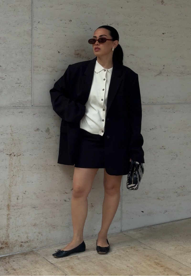 Femme portant un blazer noir et une jupe avec une chemise blanche boutonnée, des lunettes de soleil, tenant un sac à main noir et blanc à motifs devant un mur en pierre.