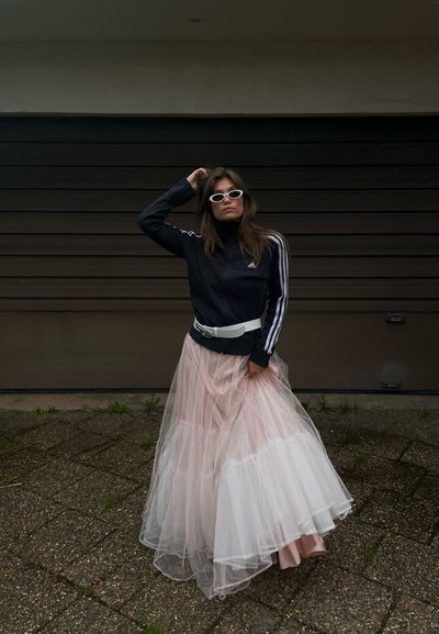 Chaqueta deportiva negra con cierre, adornada con rayas blancas, combinada con una falda de tul en varias capas en tonos rosa pálido y blanco. Los accesorios incluyen gafas de sol blancas.
