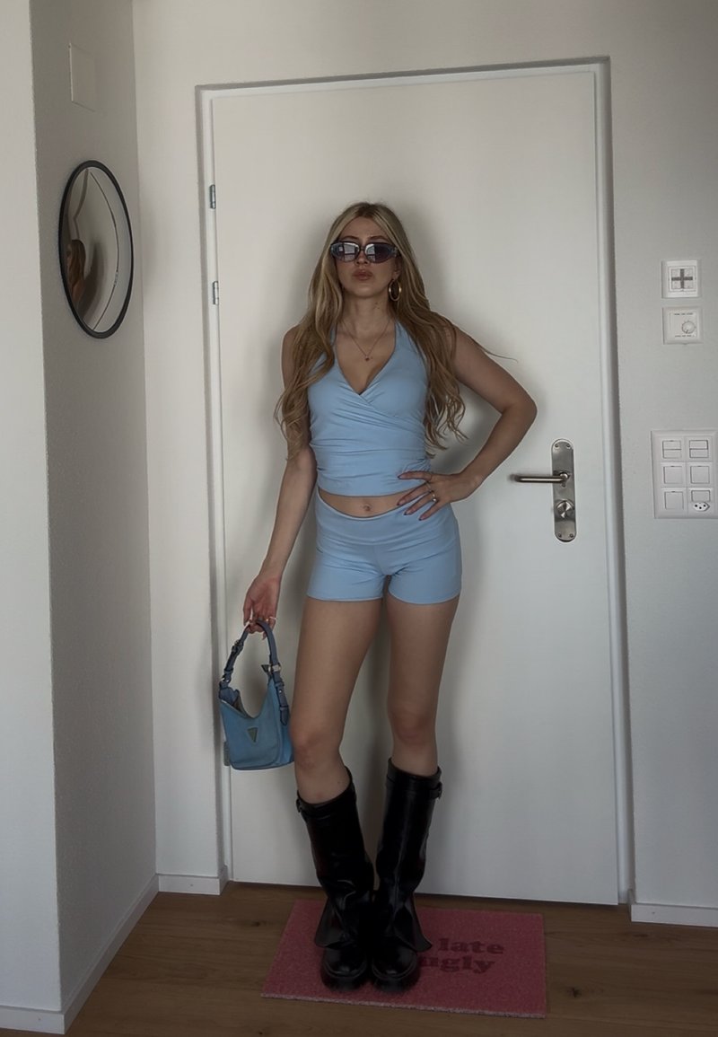 Žena s dlhými blond vlasmi, oblečená v modrom tielku bez rukávov a krátkych nohaviciach, čiernych topánkach, slnečných okuliaroch, drží modrú kabelku, stojí vo vnútri pri bielych dverách.