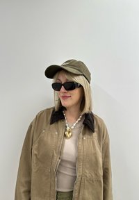 Λαδί καπέλο, μαύρα ορθογώνια γυαλιά ηλίου, μπεζ σακάκι με γιακά και καπιτονέ λεπτομέρειες, και μαργαριταρένιο κολιέ με χρυσό μενταγιόν σε σχήμα καρδιάς.