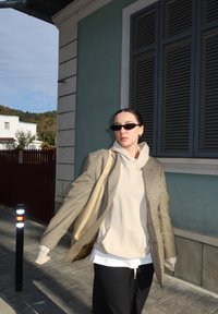 Ženska z sončnimi očali, bež kapuco, karo suknjičem in črnimi hlačami hodi na prostem blizu modre stavbe z zaprtimi roletami podnevi.