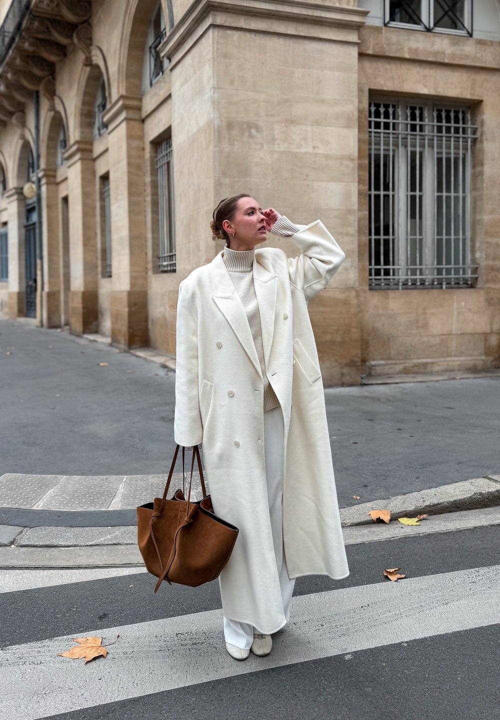 Long manteau en laine blanche à double boutonnage, associé à un col roulé crème et un pantalon à jambes larges. Sac fourre-tout en cuir marron, texture lisse.