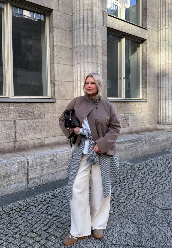 Una mujer con una chaqueta color topo y unos pantalones blancos anchos está de pie en una calle empedrada junto a un edificio de piedra y sostiene un bolso negro.