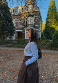 Lichtblauwe trui met een ruitpatroon in beige, gecombineerd met een lange bruine rok. Dikke donkere haren, met een bijpassende hoofdband. Herfstbladeren zichtbaar.