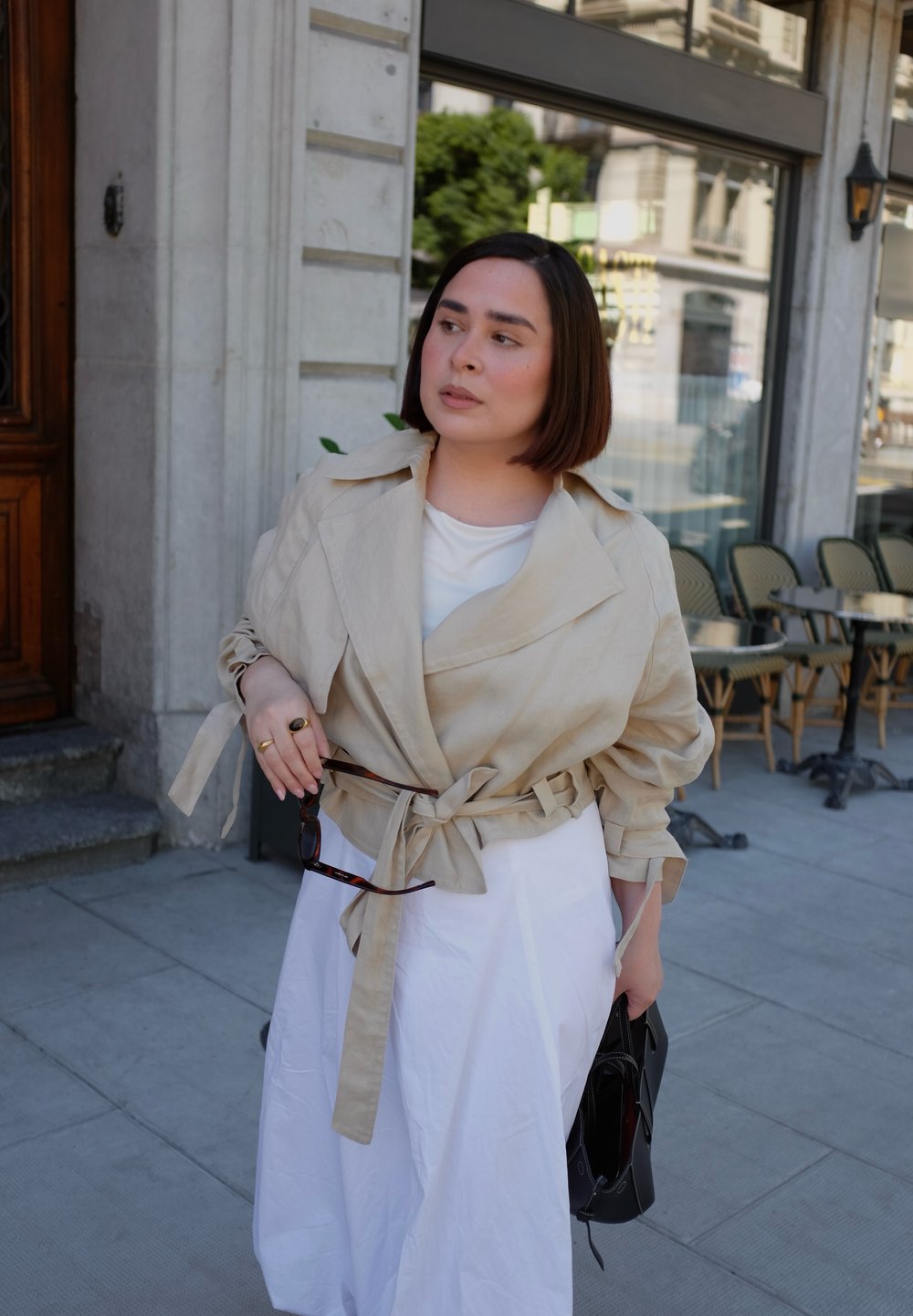 Beżowy płaszcz z lnu z dużym kołnierzem, z paskiem w talii i długimi rękawami, w połączeniu z białą sukienką i czarną torbą.