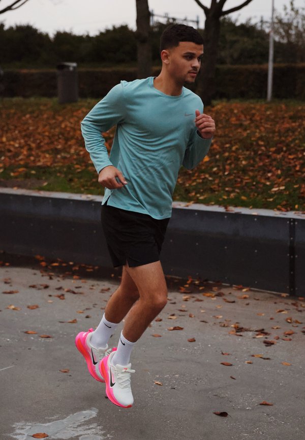 Jeune homme courant en plein air, portant un t-shirt à manches longues bleu, un short noir, des chaussettes blanches et des chaussures de course blanches avec des semelles roses sur un chemin goudronné.