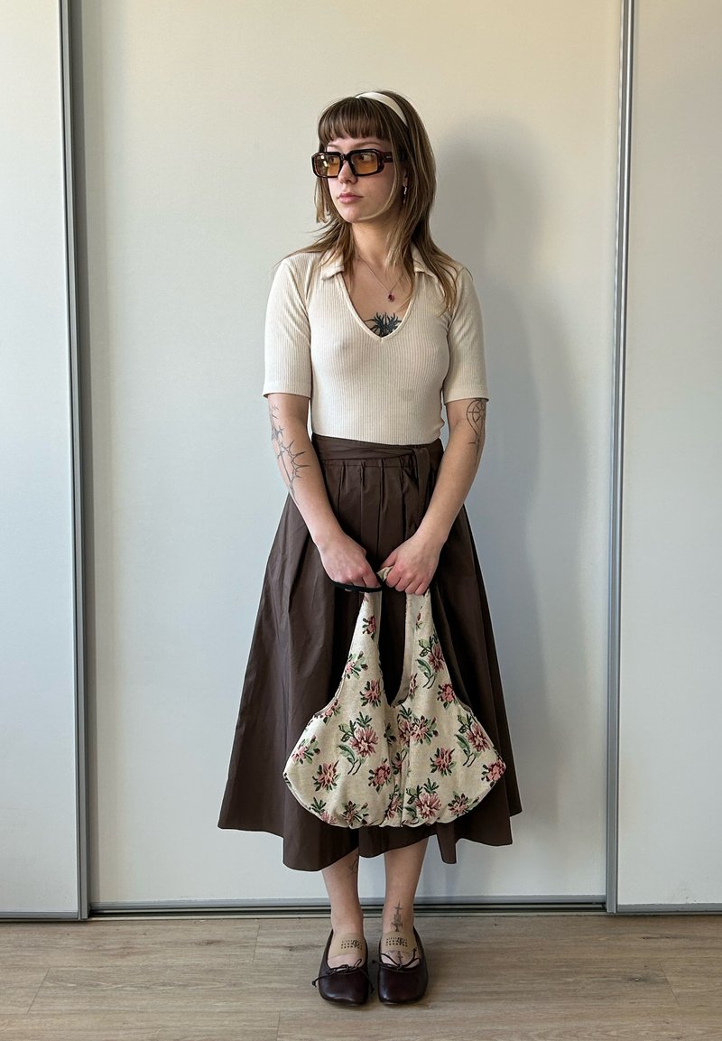 Jeune femme tatouée portant un top beige, une jupe midi marron, des chaussures marron, de grandes lunettes de soleil marron, tenant un sac à main en tissu à motifs floraux.