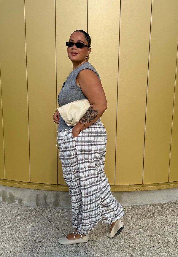 Kvinde iført sorte solbriller, grå ærmeløs top, hvide ternede bukser, beige flade sko, holder en cremefarvet clutch, stående mod en gul væg.