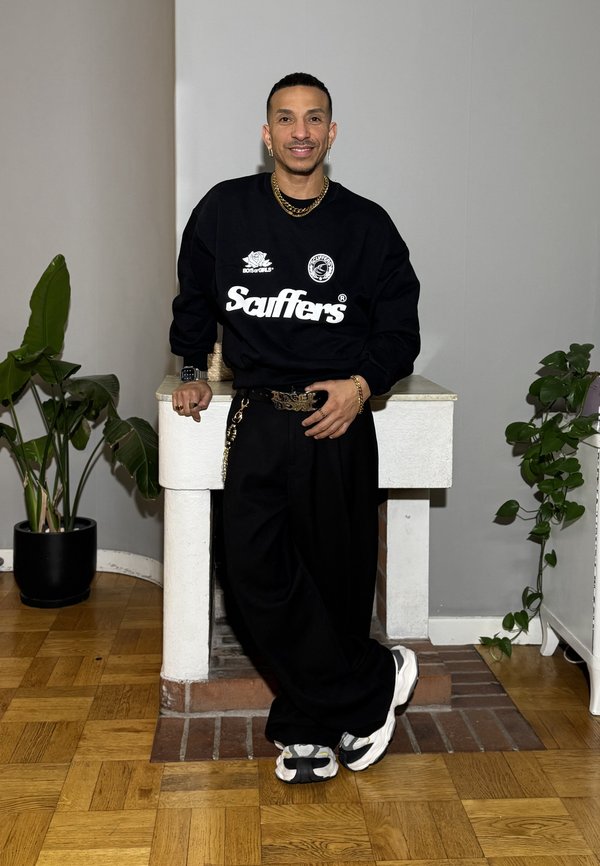 Felpa nera con logo "Scuffers", pantaloni neri larghi, gioielli argento e sneakers chunky. Ambientazione interna con piante e pareti neutre.