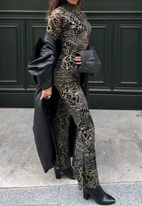 Γυναίκα που φοράει εφαρμοστό ολόσωμο ζιπ κιλτλ με μοτίβο λεοπάρδαλης, μαύρο μακρύ παλτό κρεμασμένο στους ώμους και μαύρα μποτάκια με τακούνι, στέκεται πλαγίως.