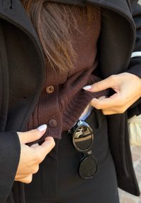 Des mains ajustant un pull marron à boutons par-dessus un pantalon noir, avec des lunettes de soleil noires accrochées à la ceinture, en plein air.
