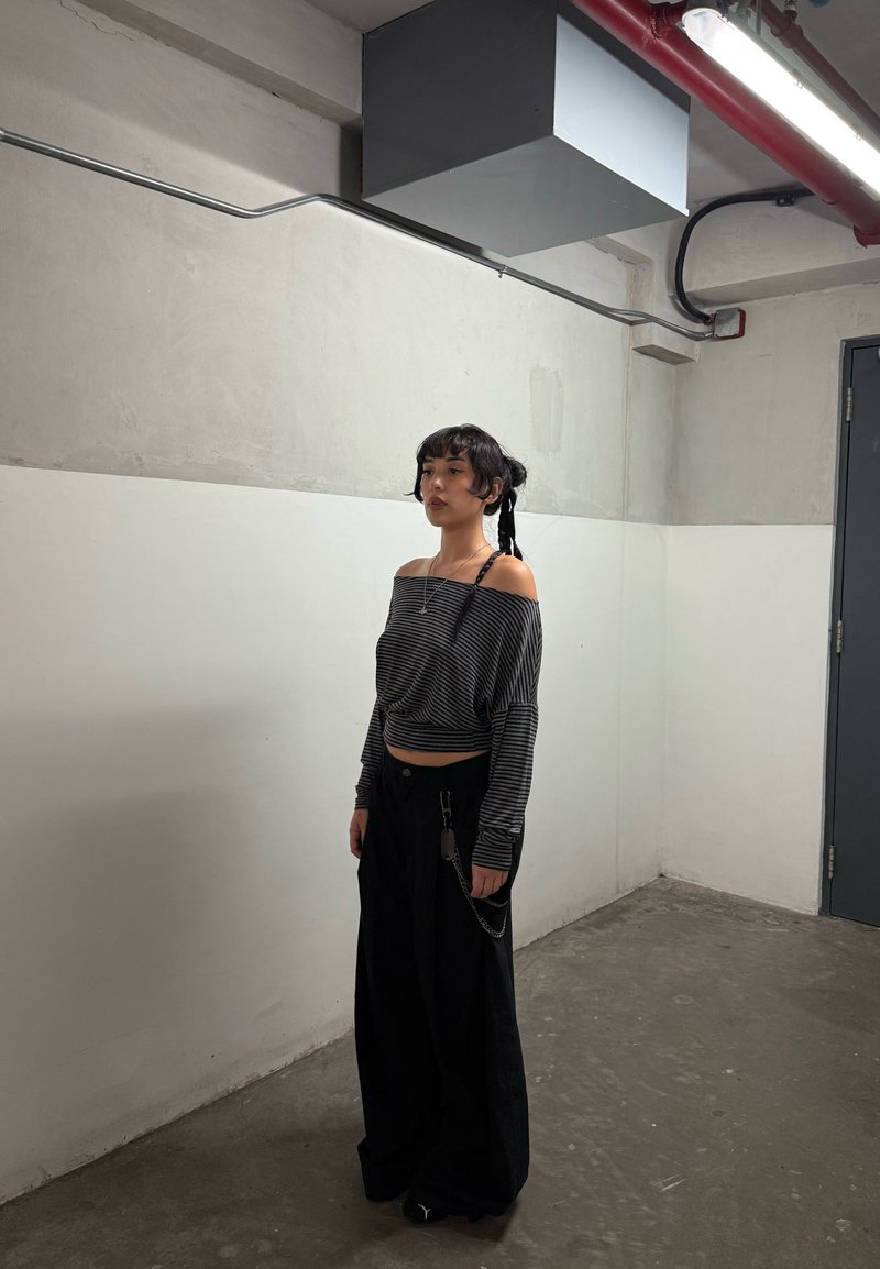 Jovem mulher com cabelo trançado vestindo uma blusa de listras com ombros à mostra e calças largas pretas, de pé numa sala industrial com paredes de concreto.