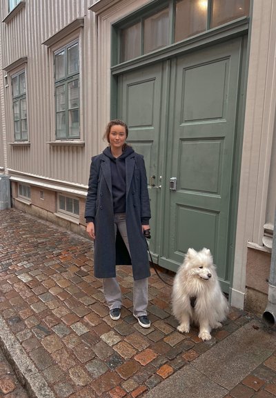 Una mujer con un abrigo oscuro y jeans grises está de pie junto a un perro Samoyedo esponjoso y blanco en una calle de adoquines, con puertas verdes al fondo.