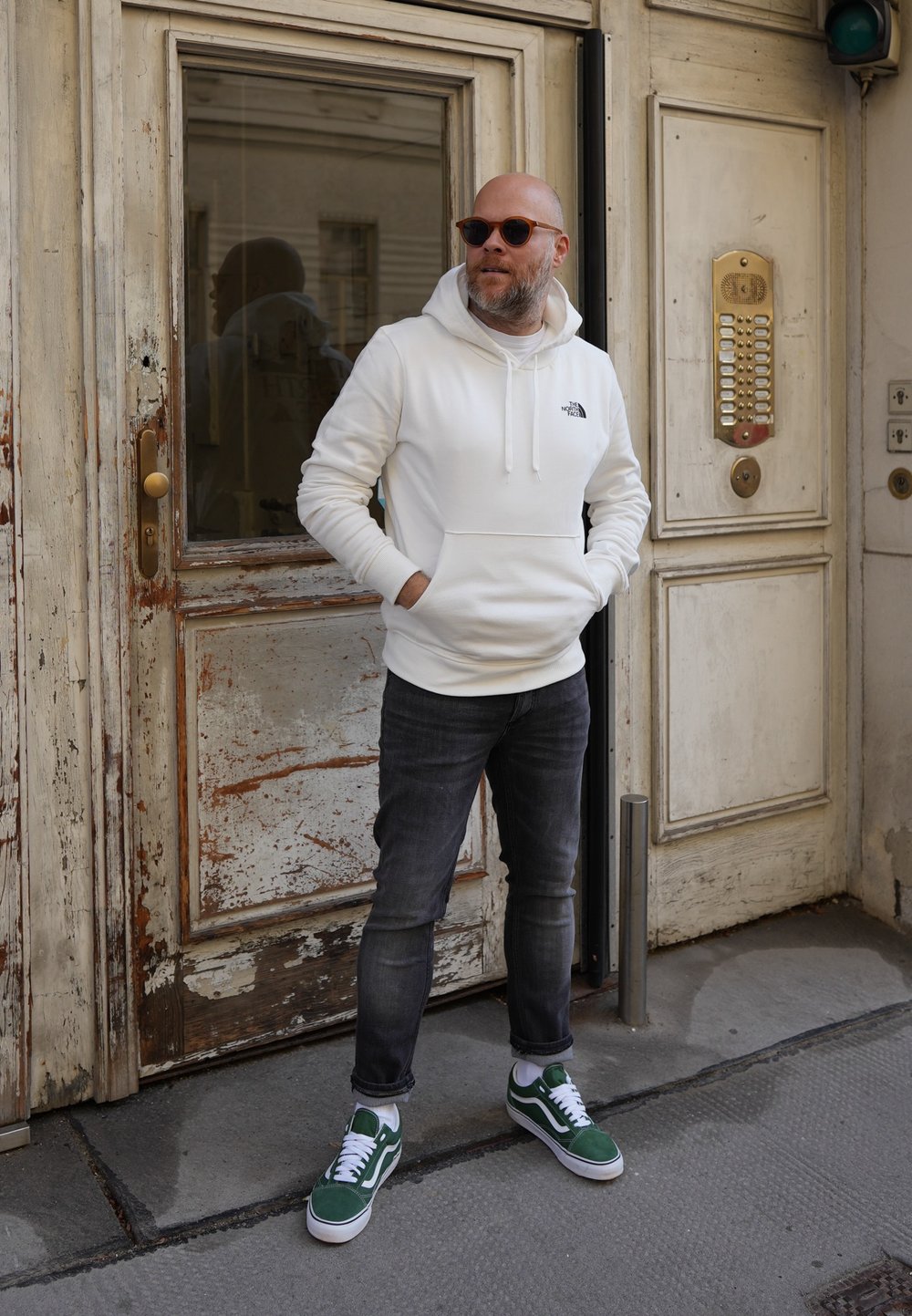 Felpa bianca con tasca frontale e logo, jeans scuri, sneakers verdi, in piedi accanto a una porta di legno texturizzata con pannello di vetro.