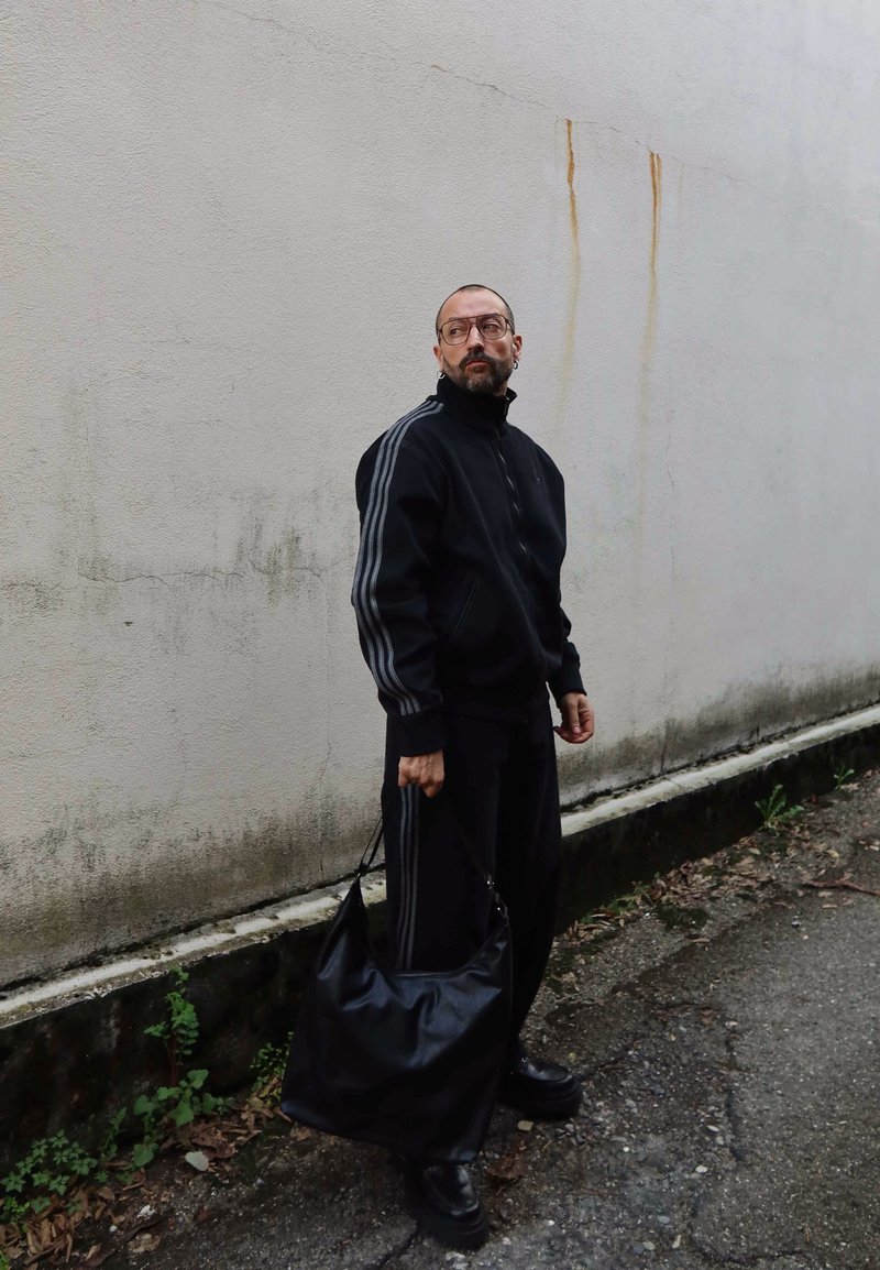 Homme avec lunettes et boucles d'oreilles en survêtement noir tenant un grand sac noir, debout contre un mur blanc usé sur un chemin extérieur.