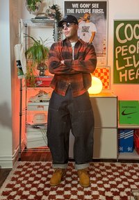 Piros és fekete kockás ing, sötét farmernadrág, barna csizmák. A kinézet napszemüveget, sapkát és laza szabást tartalmaz. A háttérben dekorációk és poszterek láthatók.