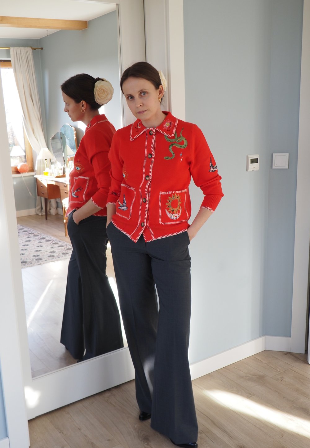 Cardigan rouge avec des motifs brodés, dont un serpent et des oiseaux, associé à un pantalon large et sombre. Sol en bois clair et miroirs reflètent la scène.