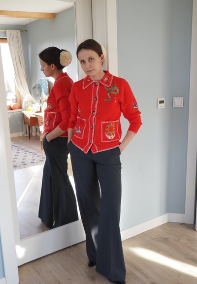 Cardigan rojo con diseños bordados, incluyendo una serpiente y pájaros, combinado con pantalones anchos de pierna oscura. Suelo de madera clara y espejos reflejan la escena.