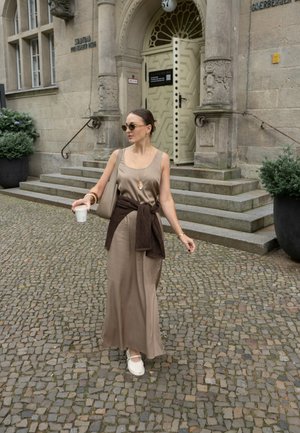 Vestido maxi de satén marrón con un suéter marrón atado a la cintura, espadrilles blancas y un bolso beige. Con un edificio de piedra de fondo.