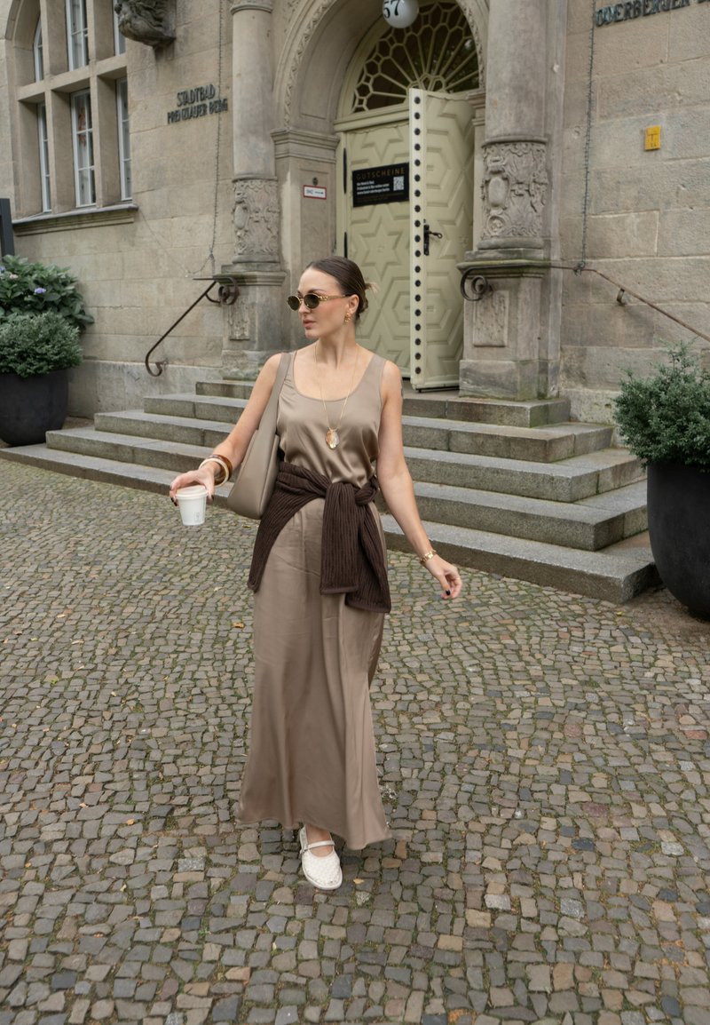 Vestido maxi de cetim castanho com um suéter castanho atado à cintura, espadrilhas brancas e uma bolsa bege. Em frente a um edifício de pedra.