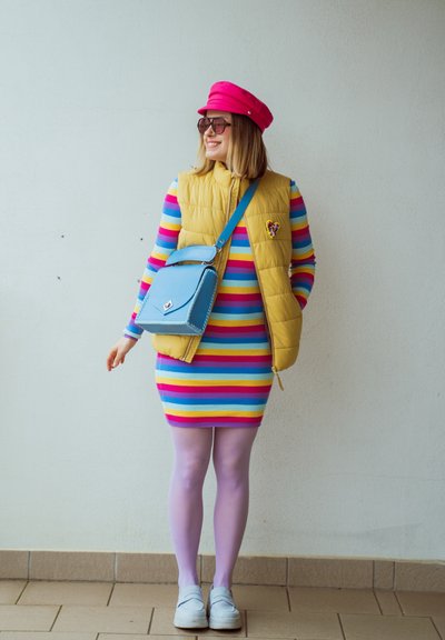 Mujer con un vestido de rayas coloridas, chaleco acolchado amarillo, sombrero rosa, medias moradas, zapatos blancos, gafas de sol y bolso azul.