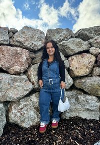 Τζιν σαλοπέτα με μπροστινό φερμουάρ, συνδυασμένη με μαύρο μακρυμάνικο πουκάμισο. Λευκή τσάντα και κόκκινα sneakers με πλατφόρμα. Φόντο με τοίχο αναρρίχησης.