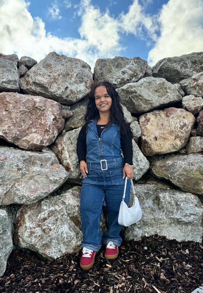 Τζιν σαλοπέτα με μπροστινό φερμουάρ, συνδυασμένη με μαύρο μακρυμάνικο πουκάμισο. Λευκή τσάντα και κόκκινα sneakers με πλατφόρμα. Φόντο με τοίχο αναρρίχησης.