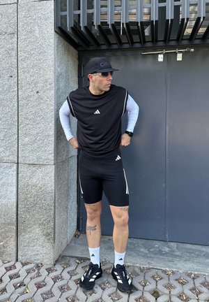 Hombre con ropa deportiva Adidas en blanco y negro, gafas de sol y gorra, de pie con las manos en la cintura junto a una puerta metálica gris y una pared de piedra.