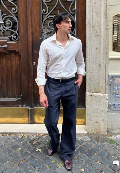 Camisa blanca de cuello, pantalones de tiro alto en color marino, cinturón negro y zapatos de cuero marrón oscuro con textura de cocodrilo; de pie junto a una puerta de madera.