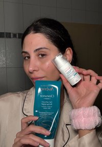 Avene reinigingsgel in een turquoise tube met witte dop, en een heldere fles Avene moisturizer, beide vastgehouden door een persoon met een roze pluche polsband.