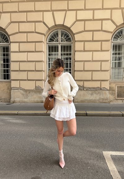 Jersey de punto blanco, falda blanca con volantes, tacones de aguja rosas y un bolso cruzado marrón. Texturas suaves y detalles en capas.