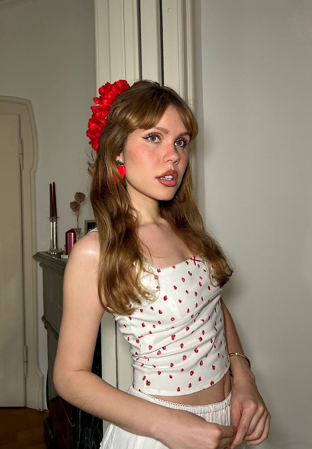 Top bianco aderente con piccoli motivi a cuore rossi, abbinato a una gonna bianca. Capelli lunghi con un accento di fiore rosso; orecchini a forma di fragola verdi.