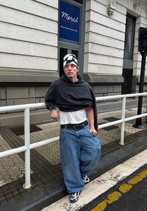 Egy szürke oversized kapucnis pulcsit, fehér pólót, laza kék farmernadrágot és mintás fekete-fehér sapkát viselő személy támaszkodik egy korlátra.