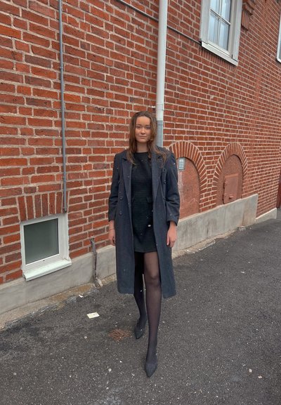 Abrigo largo negro sobre un vestido negro, mallas negras transparentes y tacones de punta. Ambientado contra un muro de ladrillos en un entorno urbano.