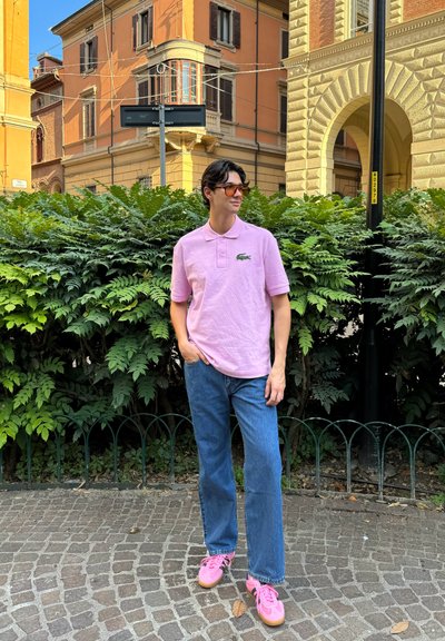 Camisa polo rosa con patrón texturizado, jeans rectos azules y zapatillas rosas con acentos negros, de pie contra un fondo de vegetación y urbano.