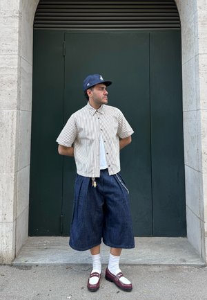 Striped short-sleeve shirt, dark denim wide shorts with pleats, white socks, and burgundy and white shoes, standing against a green door.
