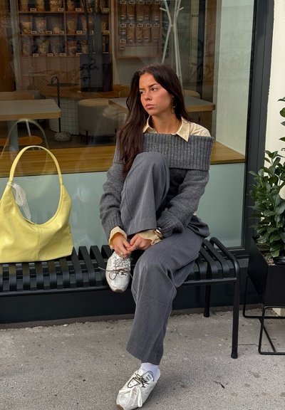 Suéter gris acanalado sobre una camisa crema, pantalones grises de pierna ancha y zapatillas blancas. Un bolso de cuero amarillo descansa sobre un banco a rayas negras.