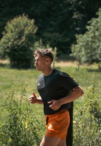 Chemise noire à manches courtes avec logo blanc, shorts orange, courant en plein air, entouré de feuillage vert et de douceur de lumière.