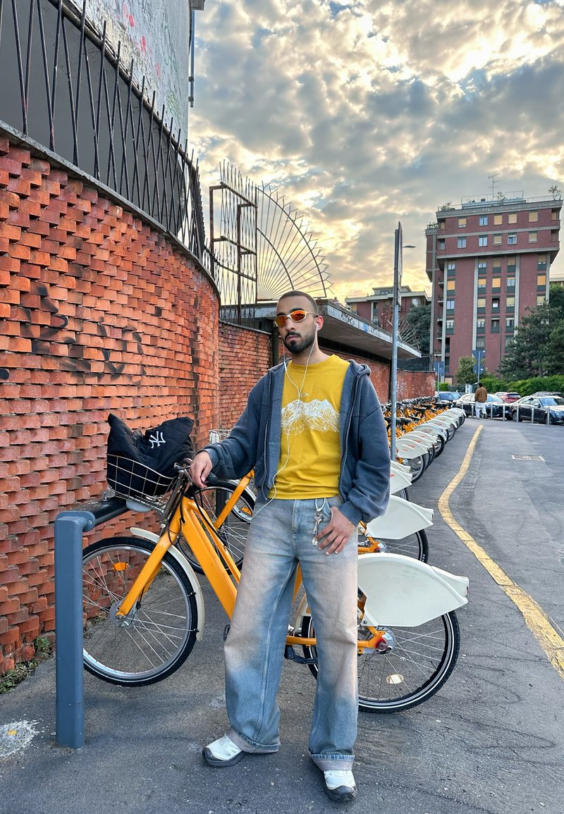 Homme portant des lunettes de soleil et des écouteurs se tient à côté d'un vélo jaune de location avec un mur de briques et un ciel nuageux en arrière-plan.