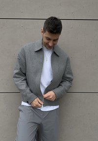 Grey wool bomber jacket with a zip front, wide collar, and cuff details, worn over a white layered shirt, paired with grey trousers.