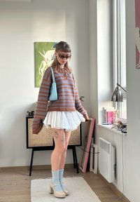 Stripet brun og blå oversized sweater, hvid flæset nederdel, lyseblå håndtaske, pastelfarvede sokker, sko med blokhæl og synlig tatovering på benene.