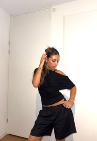 Sorte off-shoulder top med ruchet tekstur, parret med sorte shorts med høj talje. Model poserer mod en ensfarvet hvid væg.