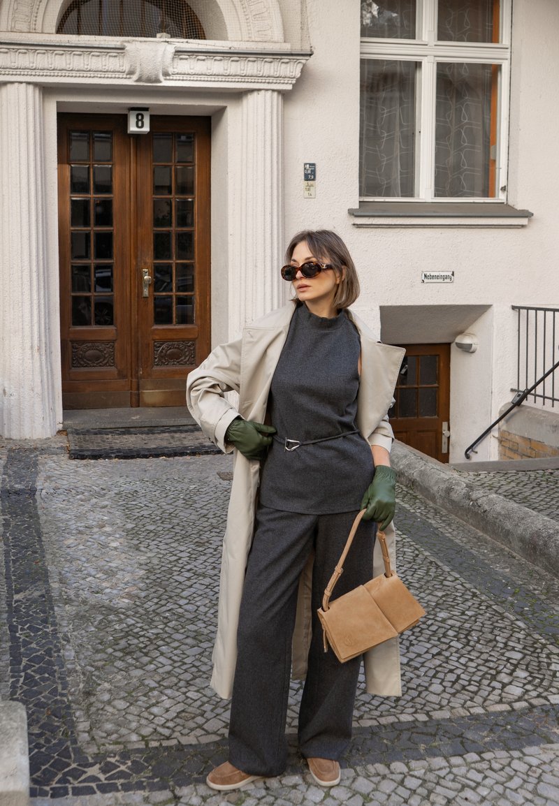 Frau in beigem Trenchcoat, dunkelgrauem Outfit, grünen Handschuhen, Sonnenbrille, mit beigefarbenem Handtasche, stehend auf einer Pflastersteinstraße nahe einer Holztür.