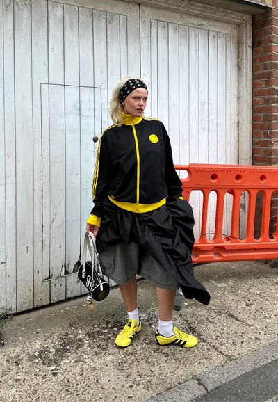 Chaqueta de chándal negra con acentos amarillos, logo sonriente amarillo, combinada con una falda negra volumétrica. Zapatillas amarillas y accesorios completan el look.