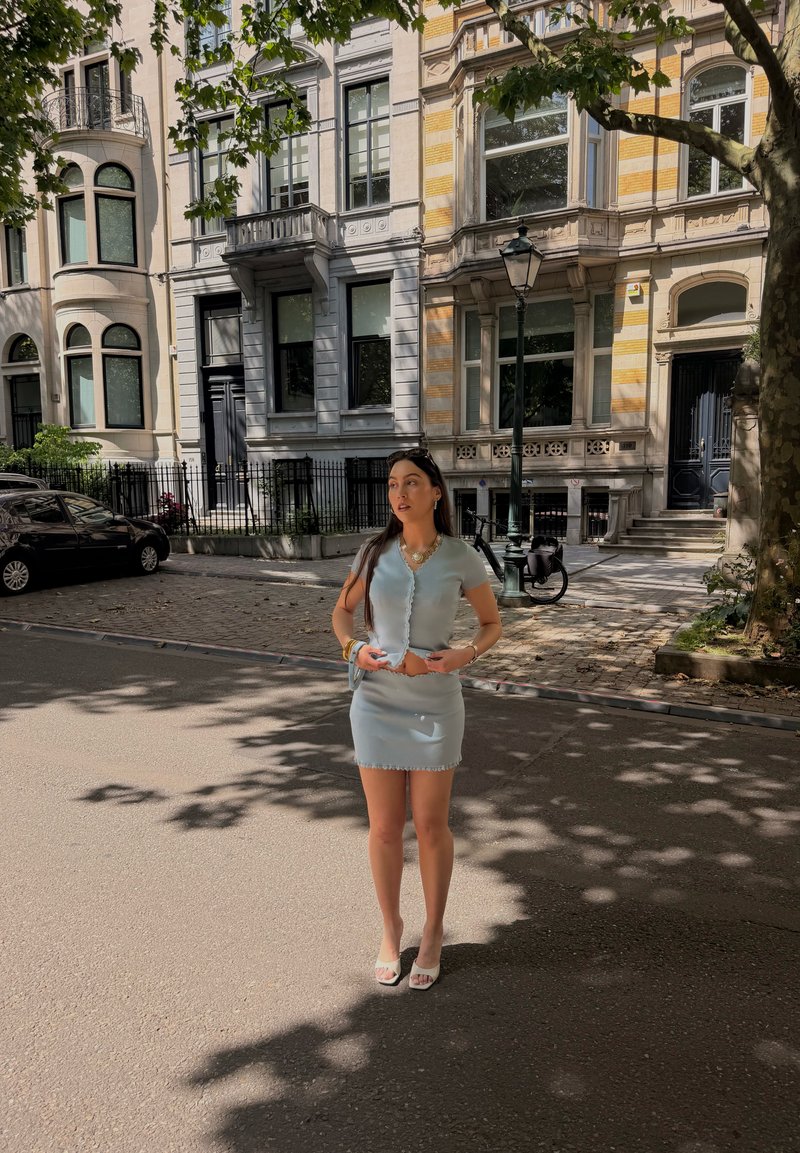 Femme portant une jupe et un haut bleu clair se tient dans une rue ensoleillée avec des arbres ombragés et des bâtiments historiques en arrière-plan.