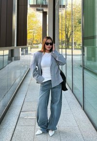 Blazer grigio oversize sopra una camicia bianca, abbinato a jeans di denim blu larghi e scarpe bianche a punta, accessoriato con occhiali da sole neri e borsa.