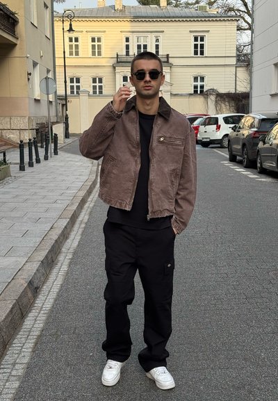 Chaqueta de pana marrón con cremallera, camiseta negra, pantalones cargo negros y zapatillas blancas. De pie en una calle de piedra.