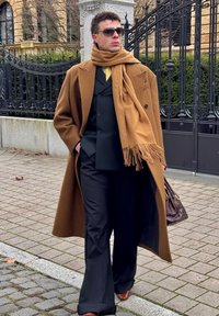 Hombre con abrigo marrón y bufanda sobre un traje negro cruzado, zapatos marrones, gafas de sol y llevando un bolso marrón oscuro con estampado.